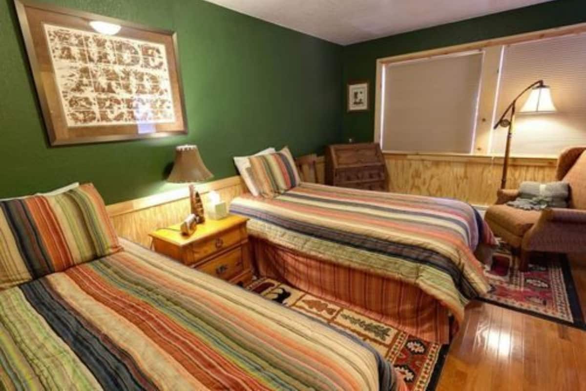 A room with two beds, a wooden headboard, a framed artwork on the wall, a lamp, and a window with blinds.