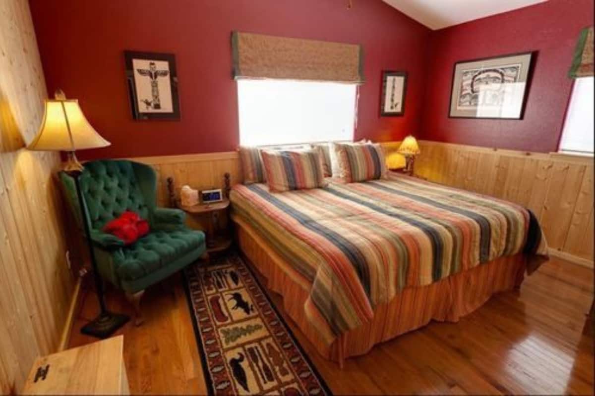 A bedroom with a bed, a green armchair, a wooden nightstand, and a window with a view.
