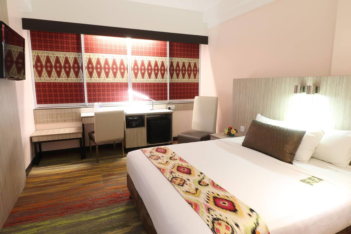A hotel room with a bed, desk, chair, and a large window with patterned curtains.