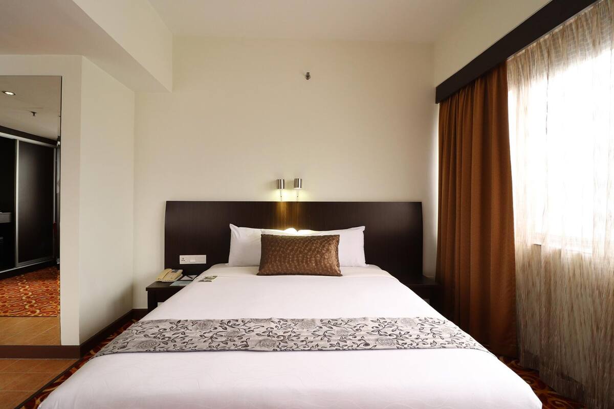 A hotel room with a large bed, a nightstand with a phone, a wall-mounted light, and a window with curtains.