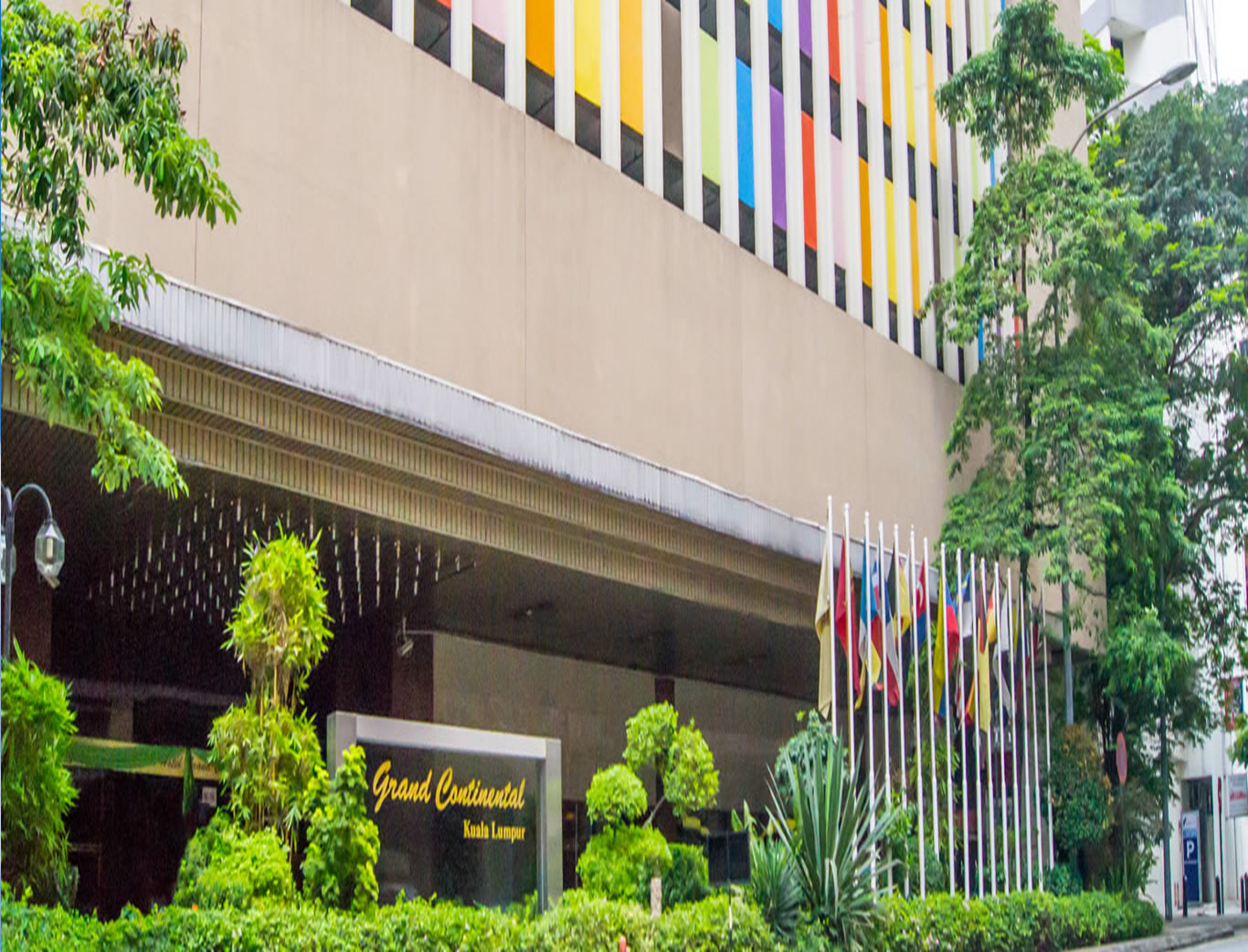Et hotel med skilt, der viser "Grand Continental Kuala Lumpur", omgivet af frodig vegetation og flere flag.