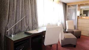 Habitación de hotel con escritorio, dos sillas, una mesita, lámpara y ventana con cortinas.