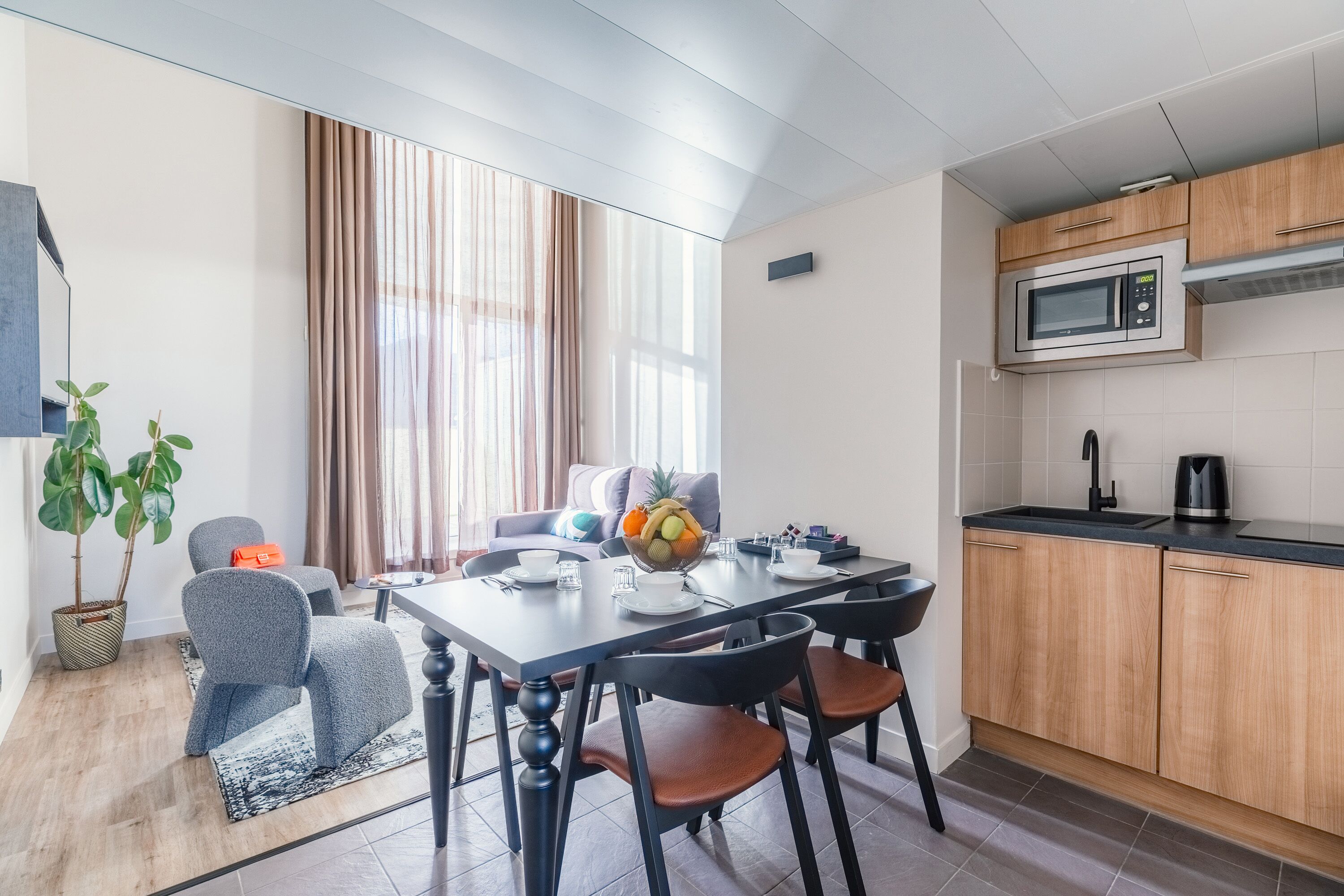 A modern kitchen with a microwave, sink, and wooden cabinets. There is a dining area with a table and chairs, and a large window with curtains.