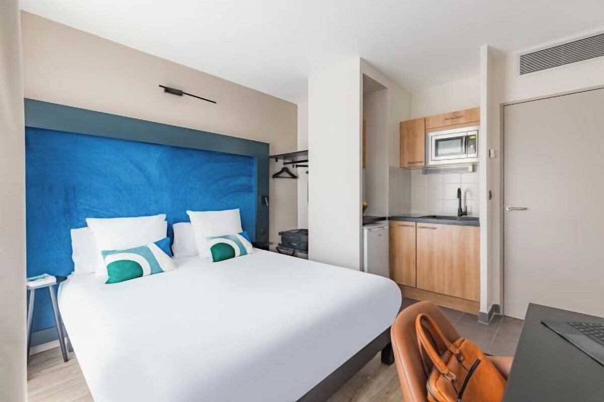 A hotel room with a bed, a desk, a chair, a microwave, and a kitchenette.
