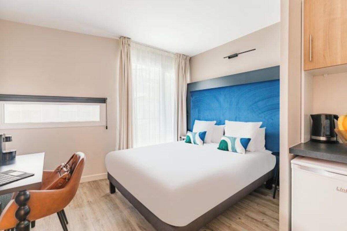 A hotel room with a bed, a desk, a chair, a kitchenette, and a window with curtains.