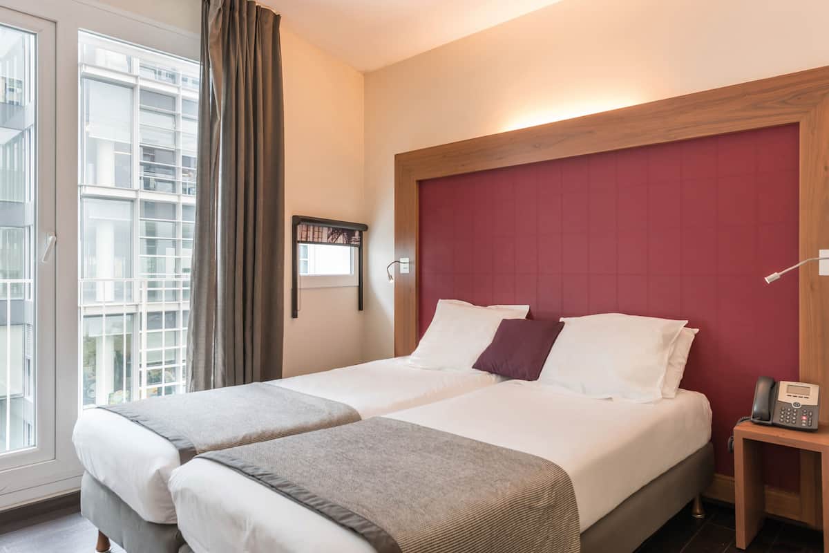 A hotel room with two beds, a red headboard, a bedside table with a phone, and a window with curtains.