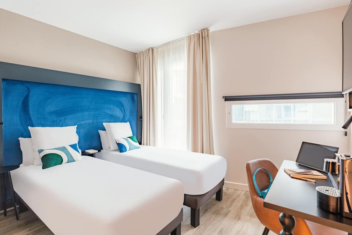 A hotel room with two beds, a desk with a laptop, and a window with curtains.