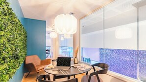 A modern meeting room with a laptop, books, and a coffee cup on a glass table, surrounded by chairs, with a green wall and a large window. - Appart'City Collection Paris Grande Bibliothèque (Paris)