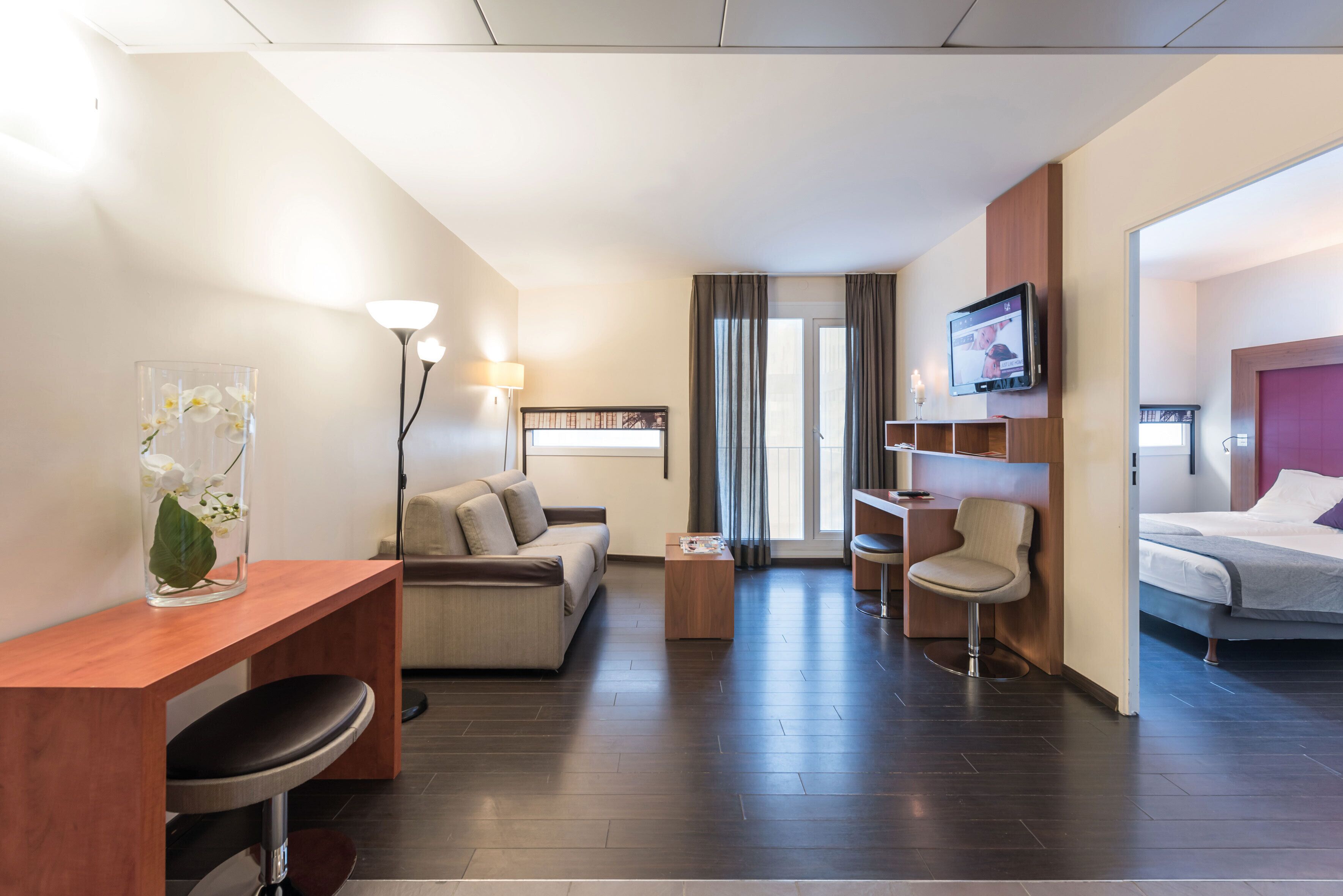 Une chambre d’hôtel équipée d’un canapé, d’un bureau, d’une chaise, d’un lit, d’une télévision et d’un vase avec des fleurs.