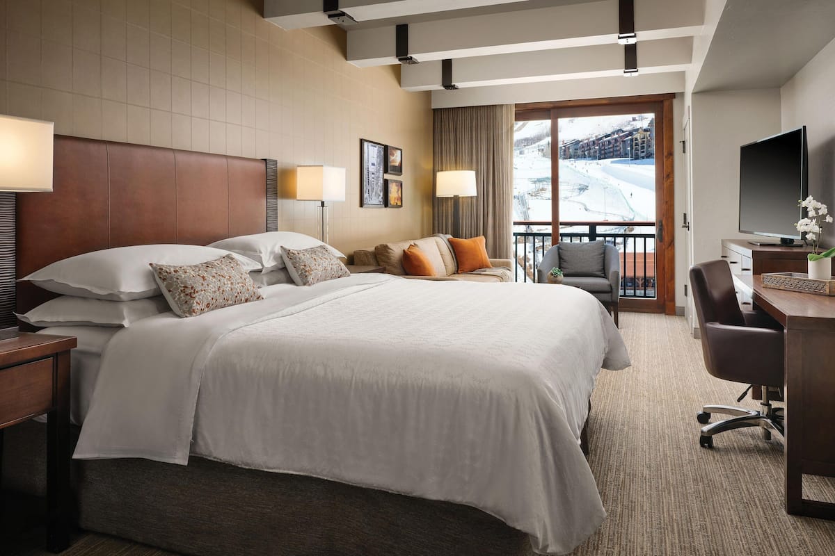 A hotel room with a large bed, a desk with a chair, a TV, and a view of a snowy landscape through the window.