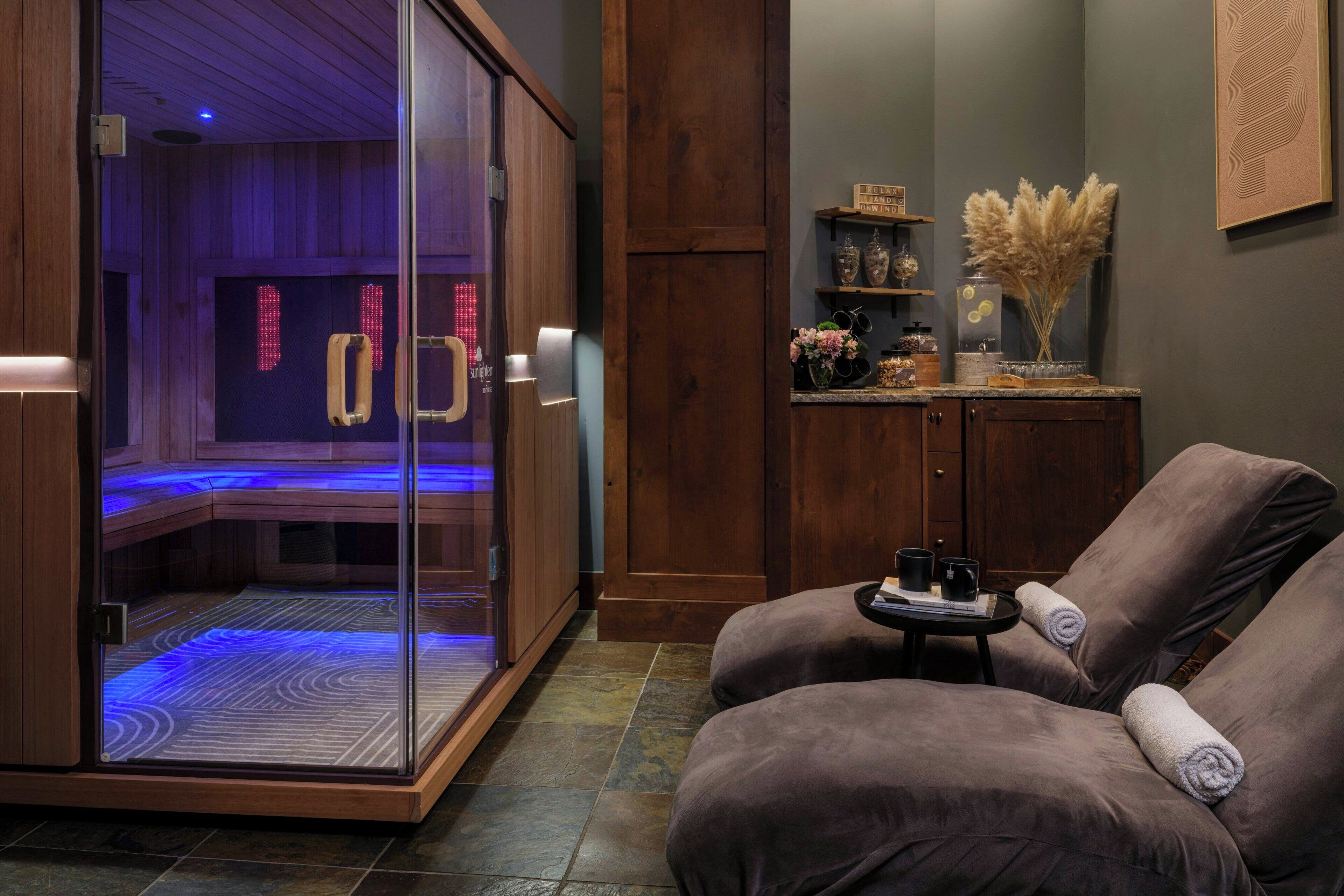 A spa room with a glass-enclosed sauna, a wooden cabinet with decorative items, and a relaxation area with cushioned chairs.