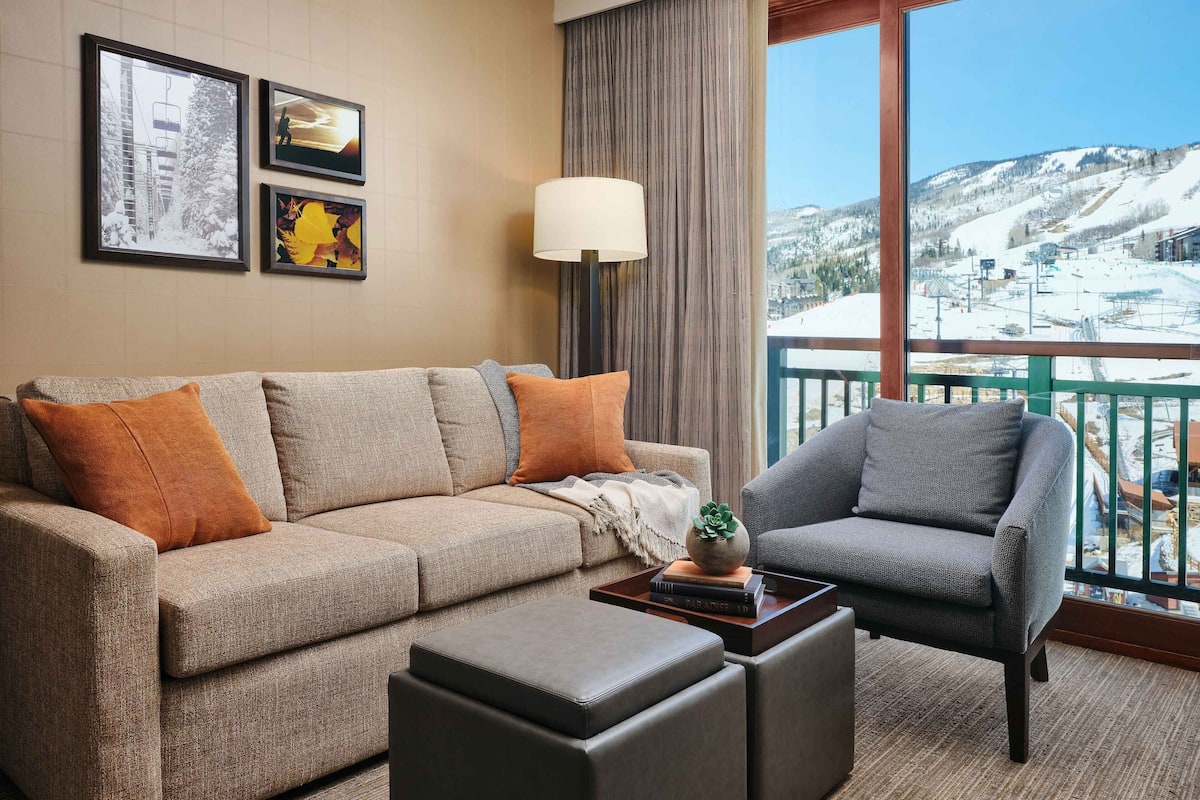 A living room with a sofa, armchair, and ottoman, a lamp, and framed pictures on the wall.