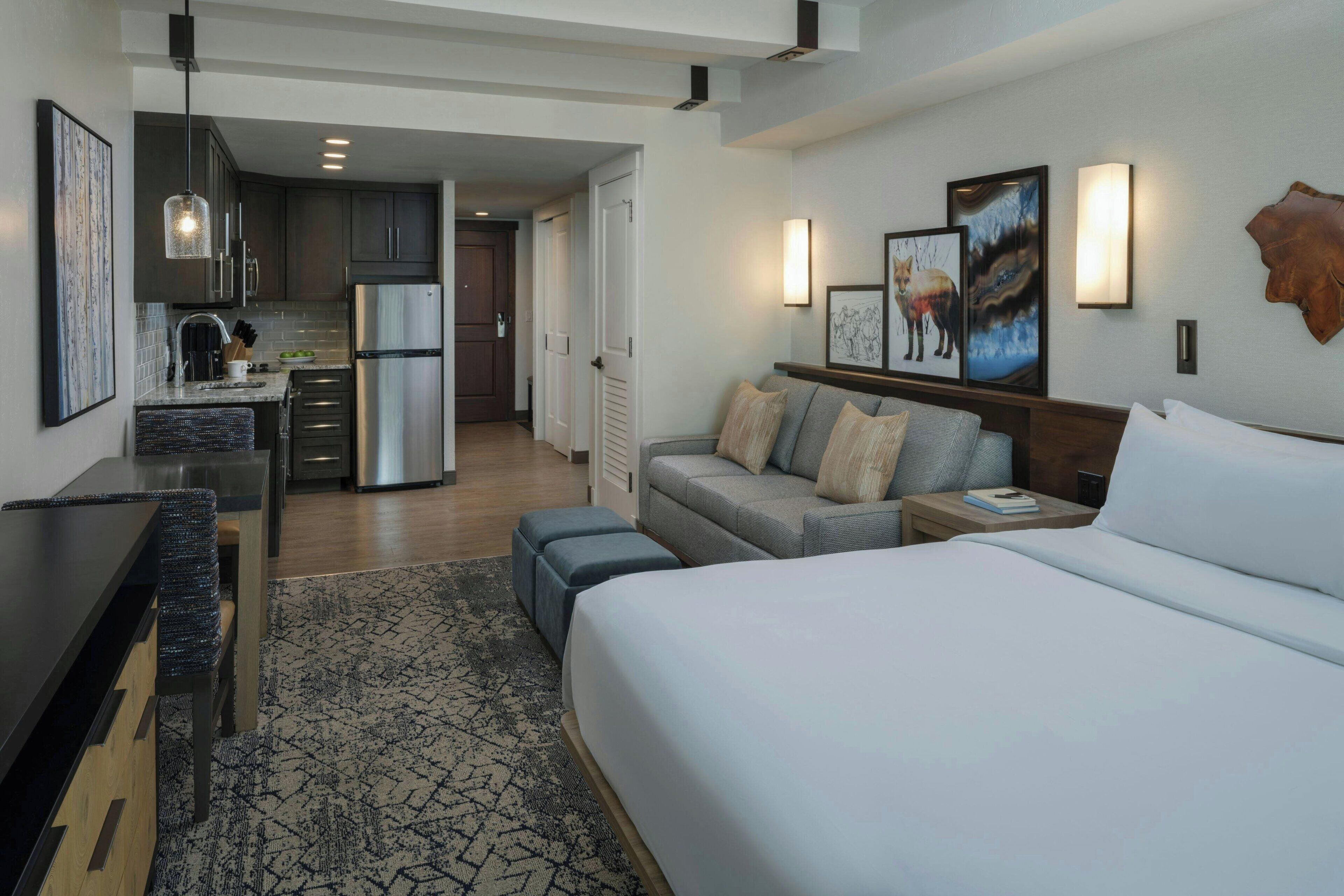 Une chambre d’hôtel moderne avec un grand lit, un canapé, un bureau et une kitchenette. Les murs sont ornés de tableaux encadrés et recouverts d’un revêtement en cuir.