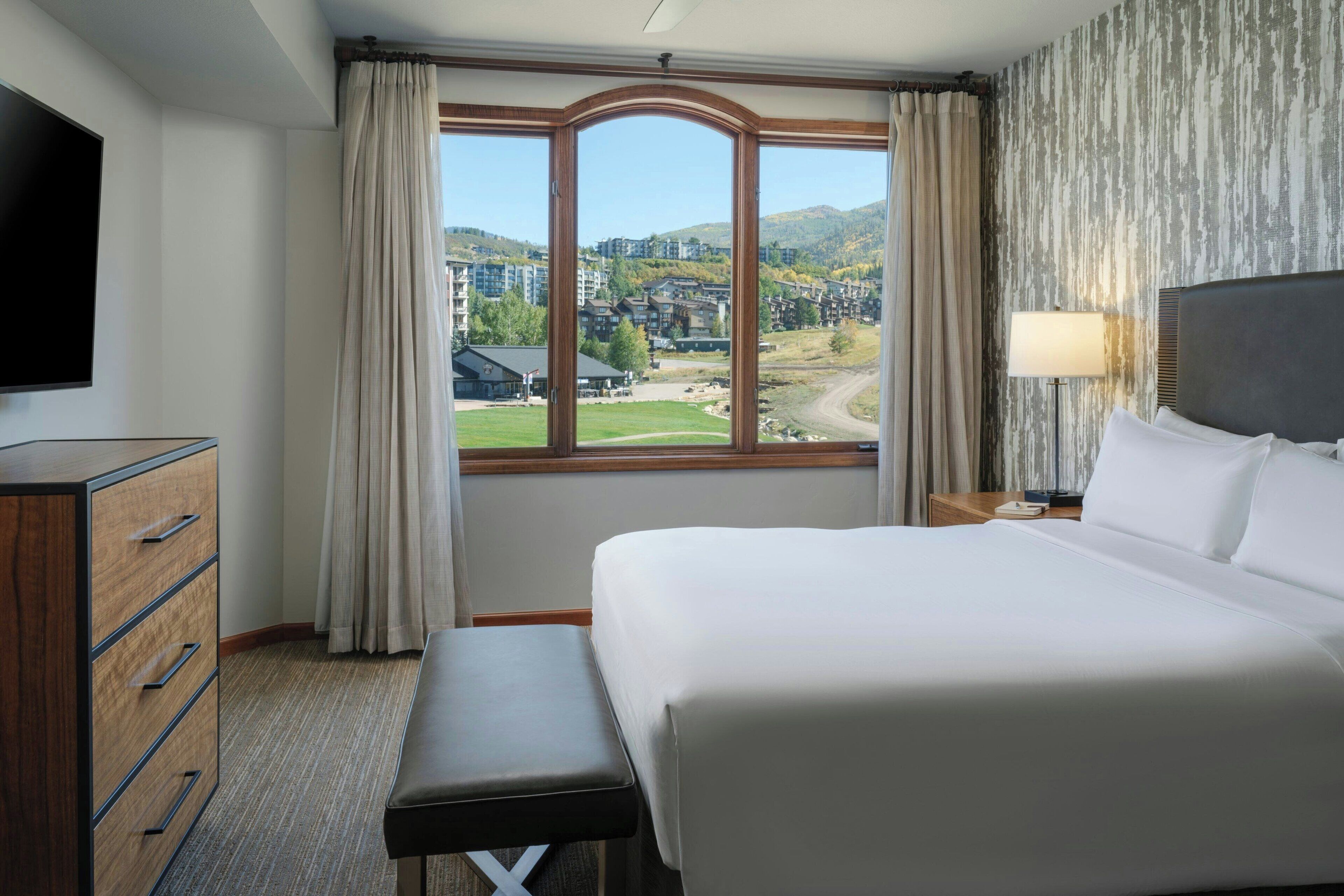 Ein Hotelzimmer mit einem großen Bett, einem Schreibtisch mit Schubladen, einem Fernseher und einem Fenster mit Blick auf Gebäude und Hügel.