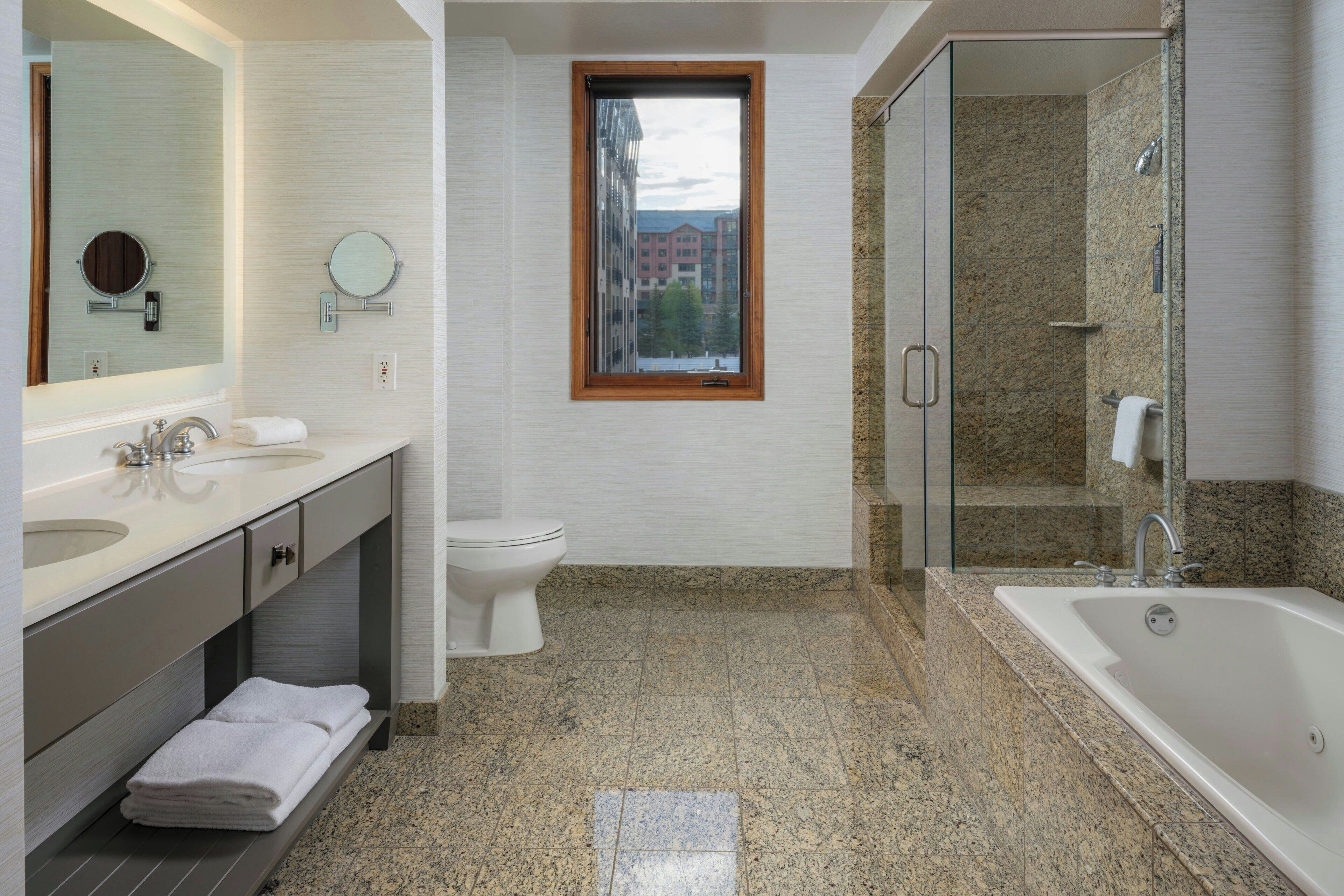 A modern bathroom with a large bathtub, a separate toilet, and a window with a view of buildings.