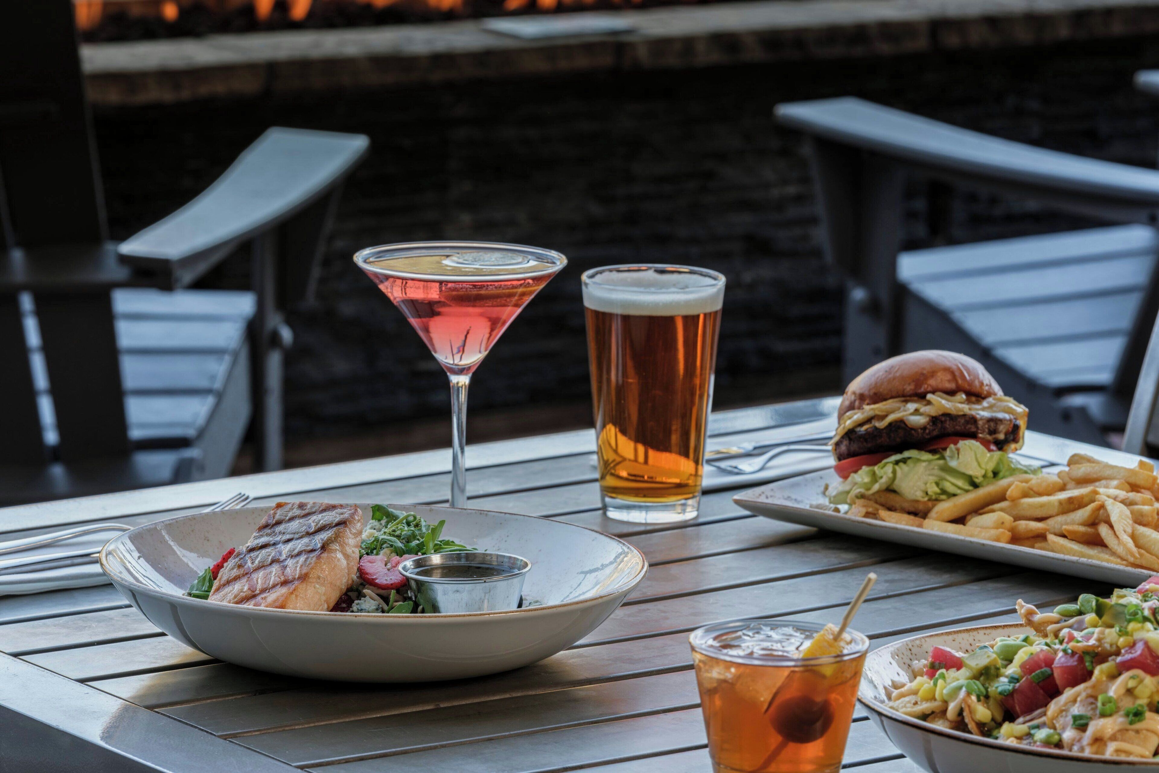A dining table with a plate of food, a martini glass, a tall beer glass, a burger, fries, and a salad.