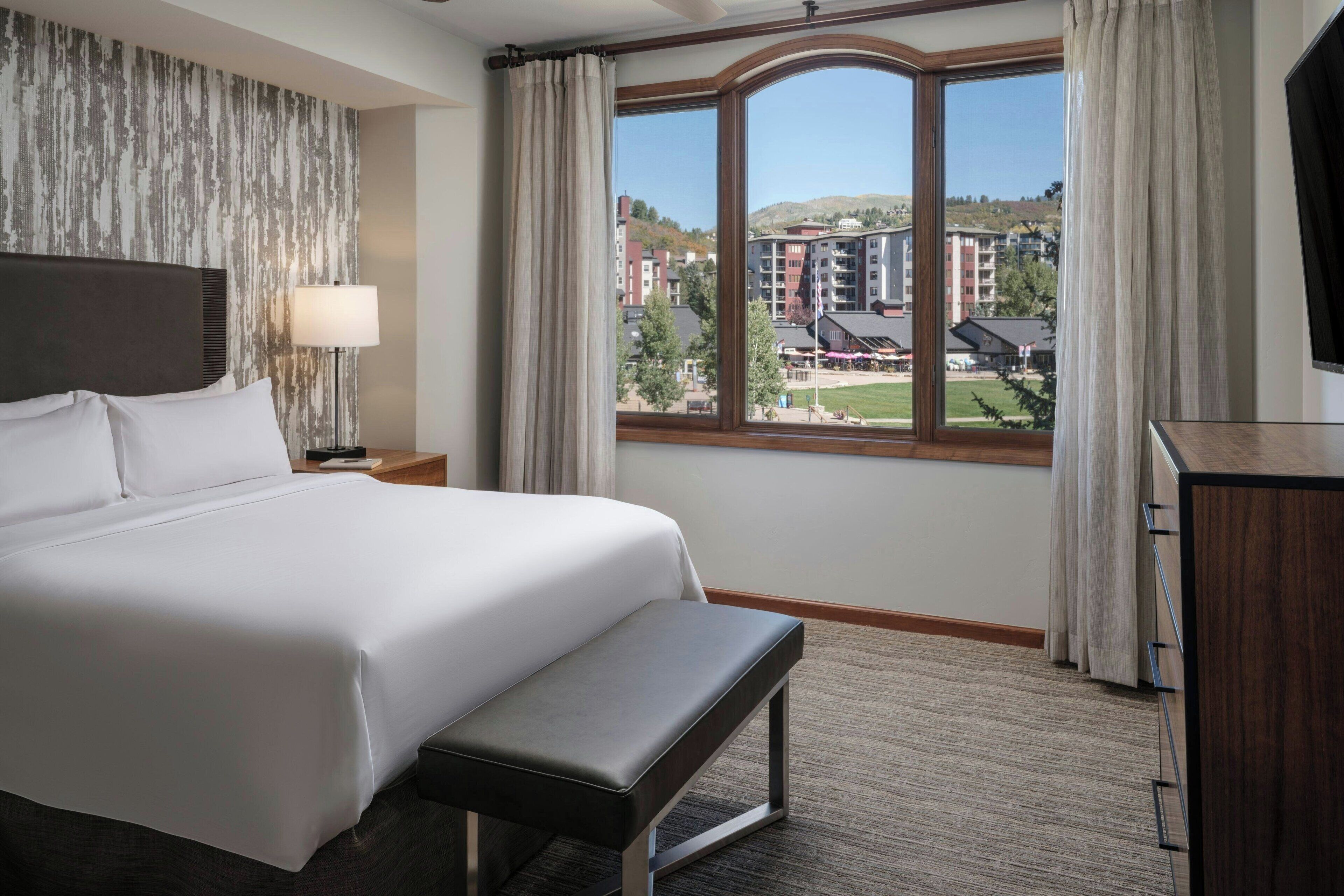 A hotel room with a large bed, a bench, a TV, and a view of buildings and a green area outside.
