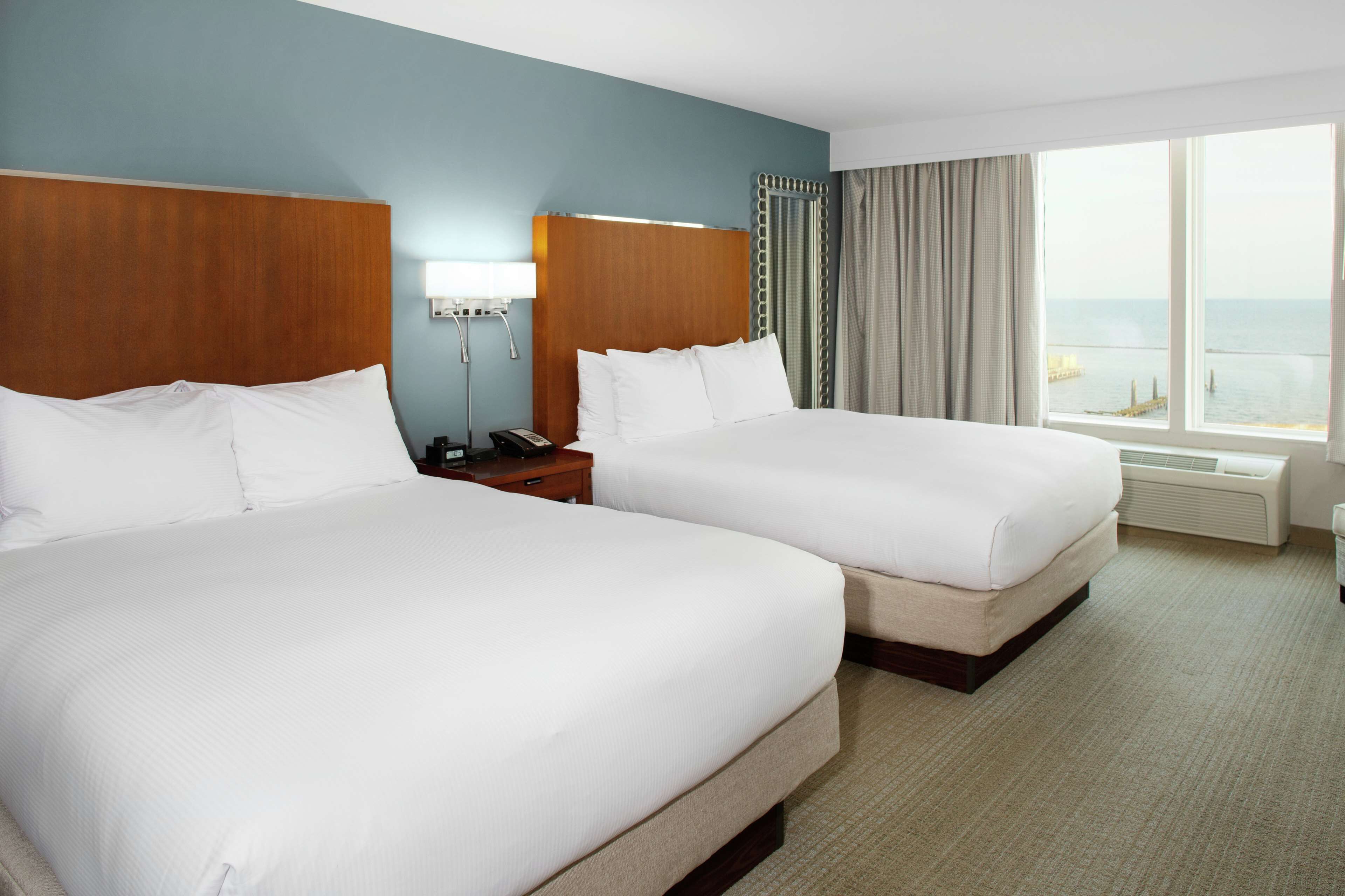 A hotel room with two beds, a nightstand, a lamp, and a large window with a view of the sea.
