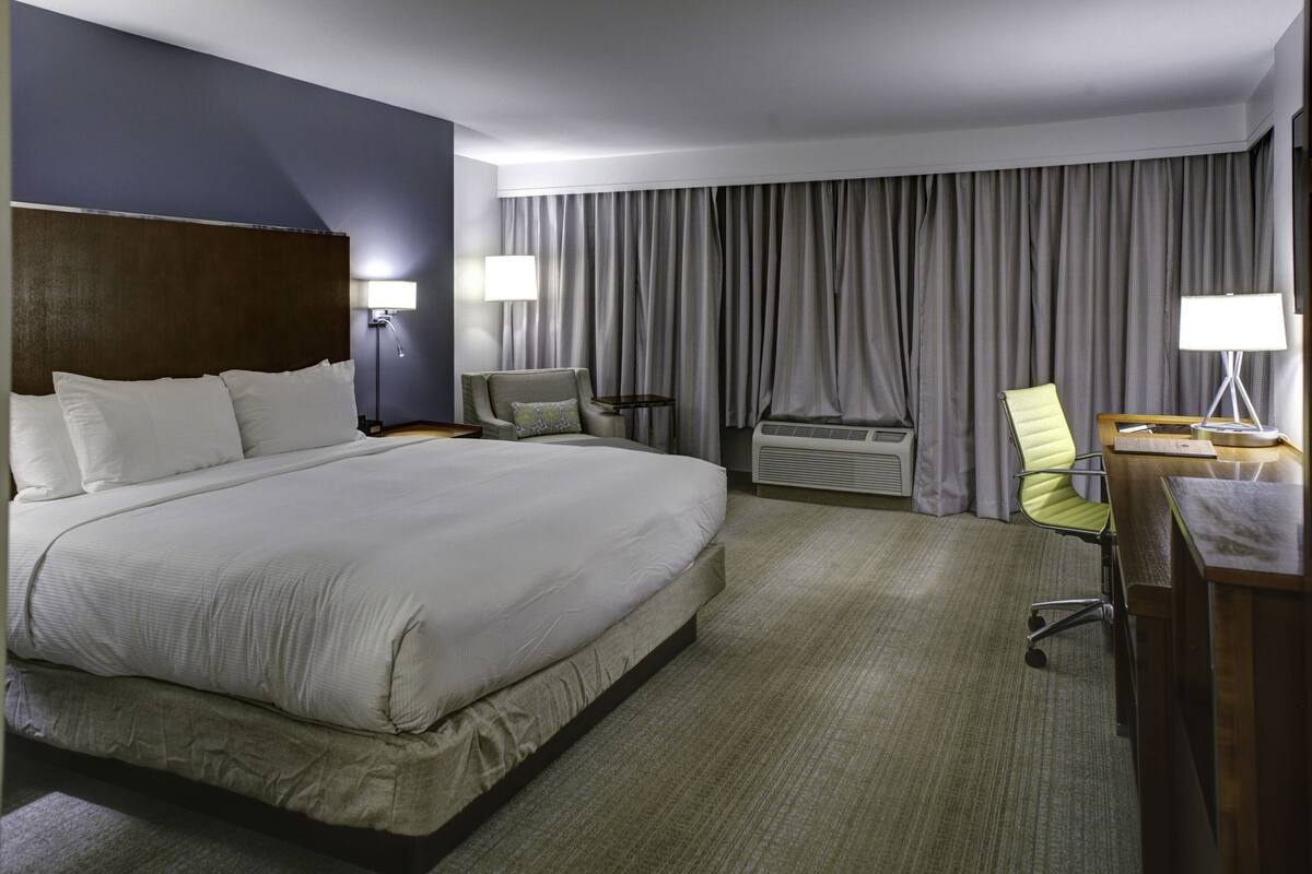 A hotel room with a large bed, a desk with a chair, a lamp, a chair, and a window with curtains.