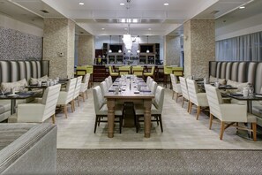 Amplia sala de comedor de hotel con varias mesas y sillas, una barra central y un suelo con diseño.