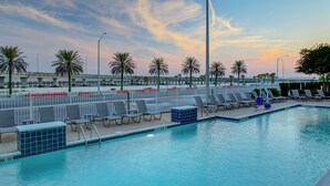 A swimming pool with lounge chairs and palm trees, surrounded by a fence and a bridge in the background. - DoubleTree by Hilton Hotel Biloxi (Biloxi)