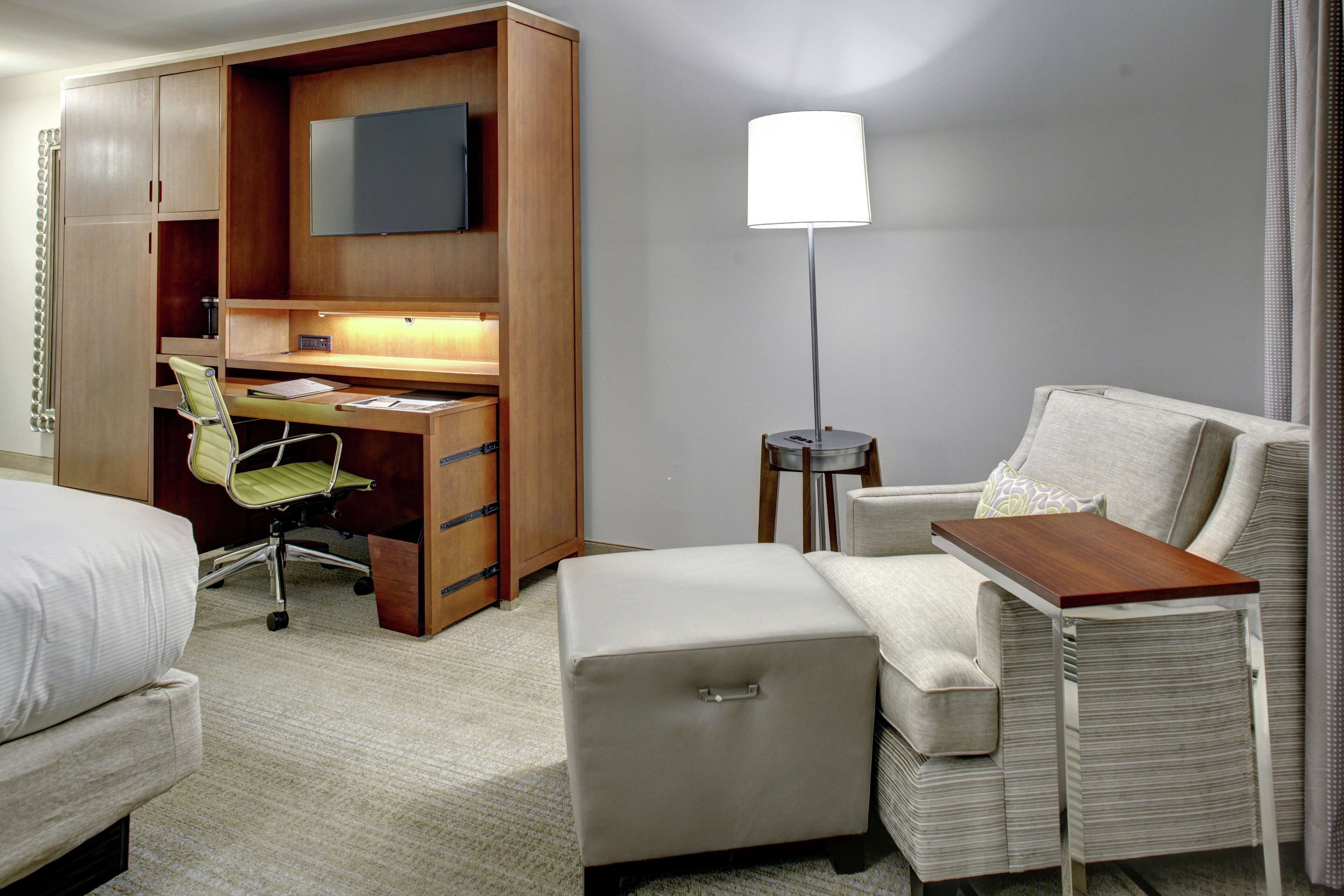 A hotel room with a bed, a desk with a chair, a small ottoman, a lamp, and a TV on the wall.