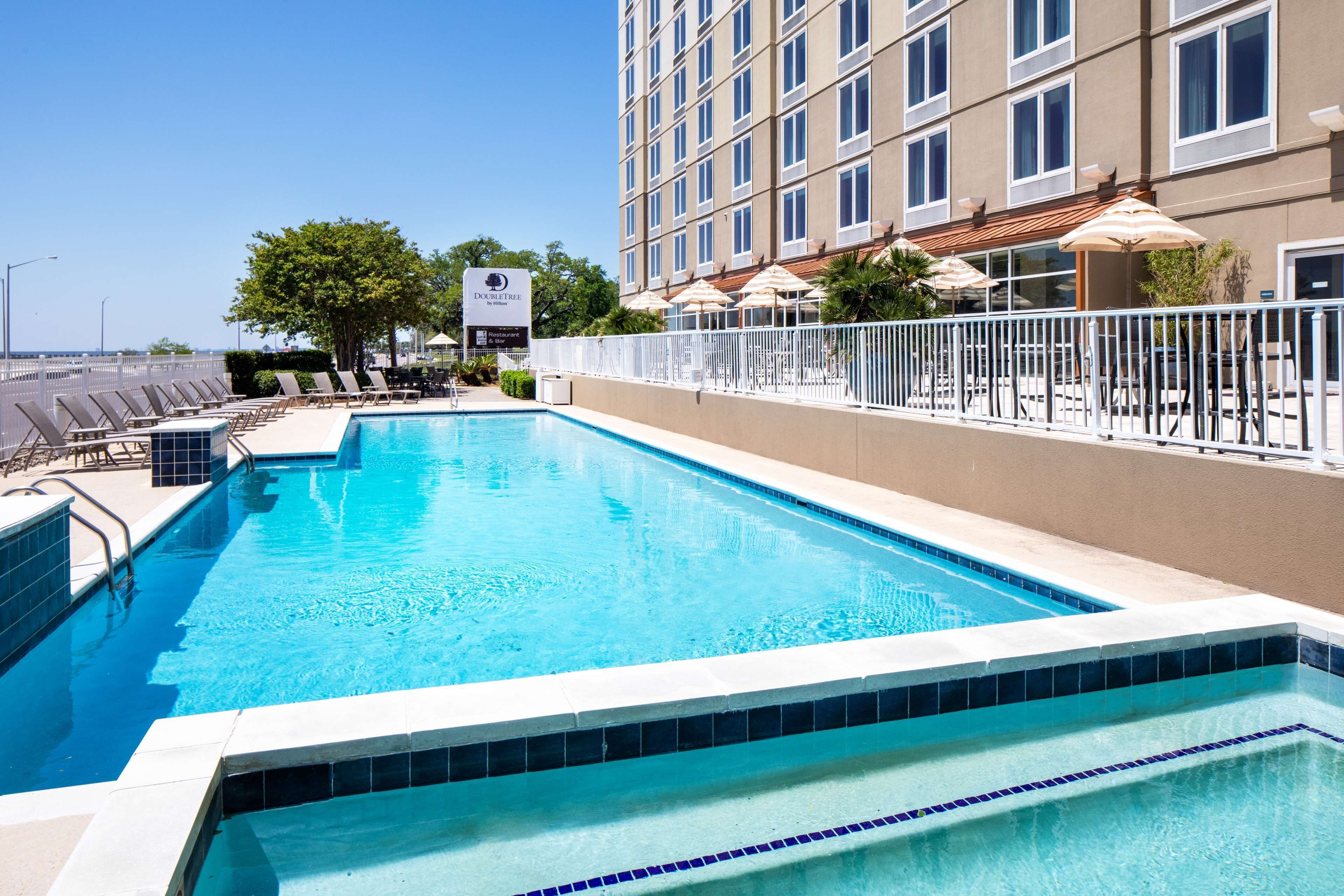 A rectangular swimming pool with a tiled edge, surrounded by a white fence, and adjacent to a building with multiple windows and outdoor seating areas.