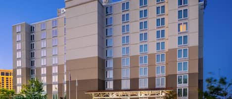 Een hotel met meerdere verdiepingen, met een modern design, een overdekte ingang en een parkeerplaats met diverse auto's.
