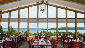 A restaurant with large windows offering a view of a lake and hills. Tables are set with red chairs. - Silver Dart Lodge (Baddeck)