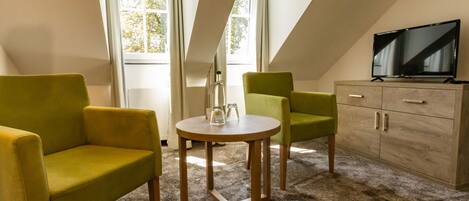 A room with two green armchairs, a small round table, a television on a cabinet, and a window with curtains.