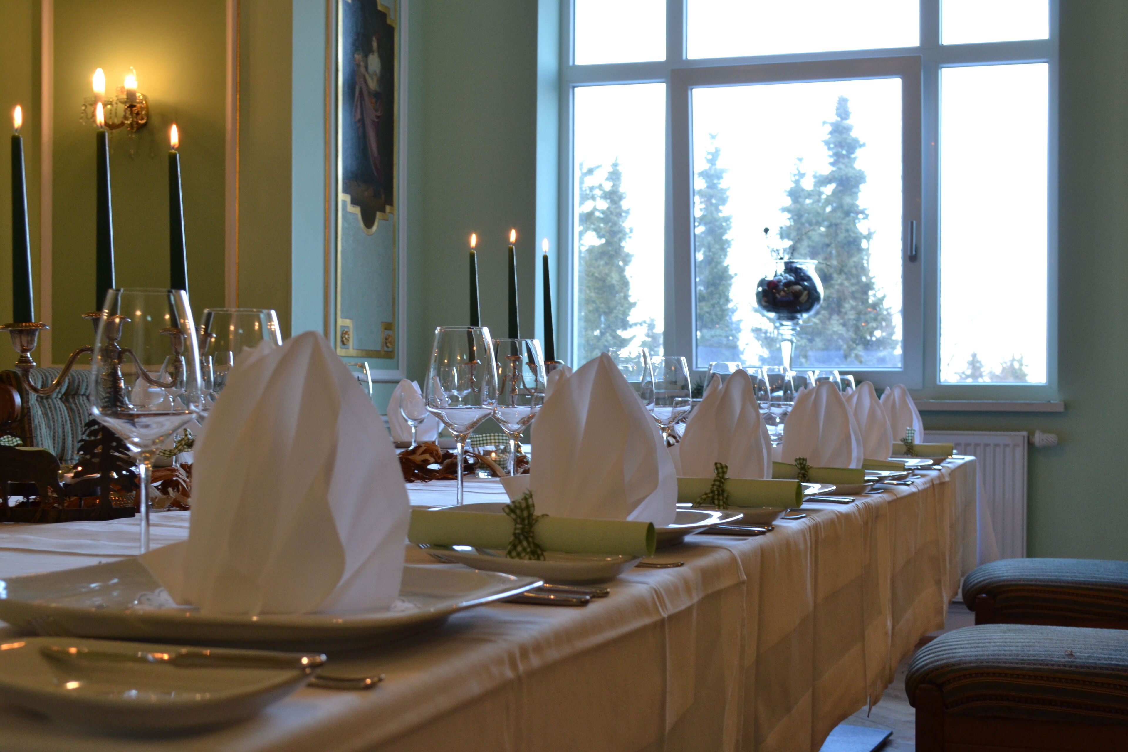 Una larga mesa de comedor dispuesta con servilletas blancas, platos, vasos, velas y vistas a árboles a través de un amplio ventanal.