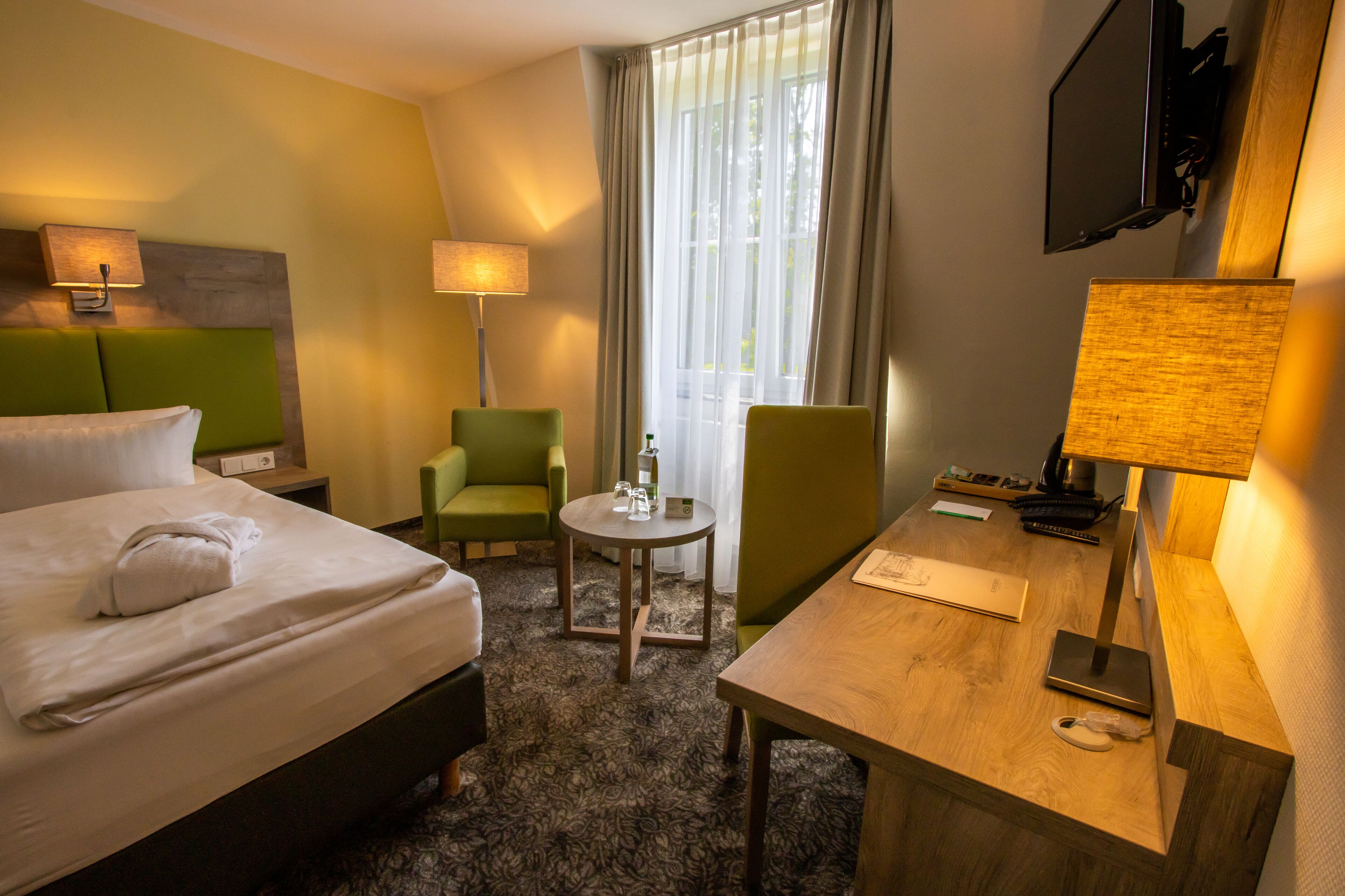 Hotellihuone, jossa on sänky, työpöytä, tuoli, lamppu ja seinälle asennettu televisio.