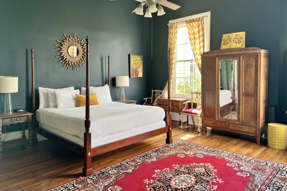 A four-poster bed with white linens, a wooden floor, a red patterned rug, a wooden wardrobe, and a ceiling fan.