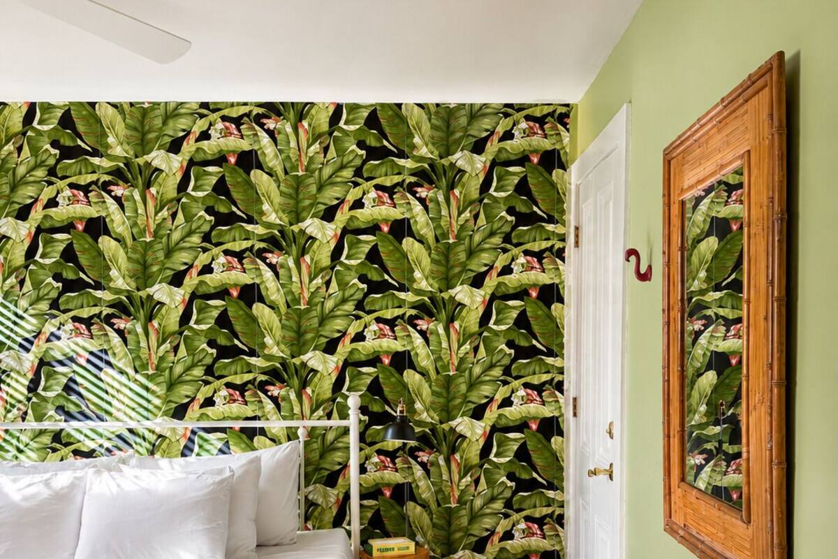 A bedroom with a bed, a wooden floor, a wall with a leafy wallpaper, a white door, and a framed picture on the wall.