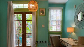 A room with a wooden desk, a chair, a hanging light fixture, a window with blinds, and a door with glass panels. - Creole Gardens Guesthouse and Inn (New Orleans)