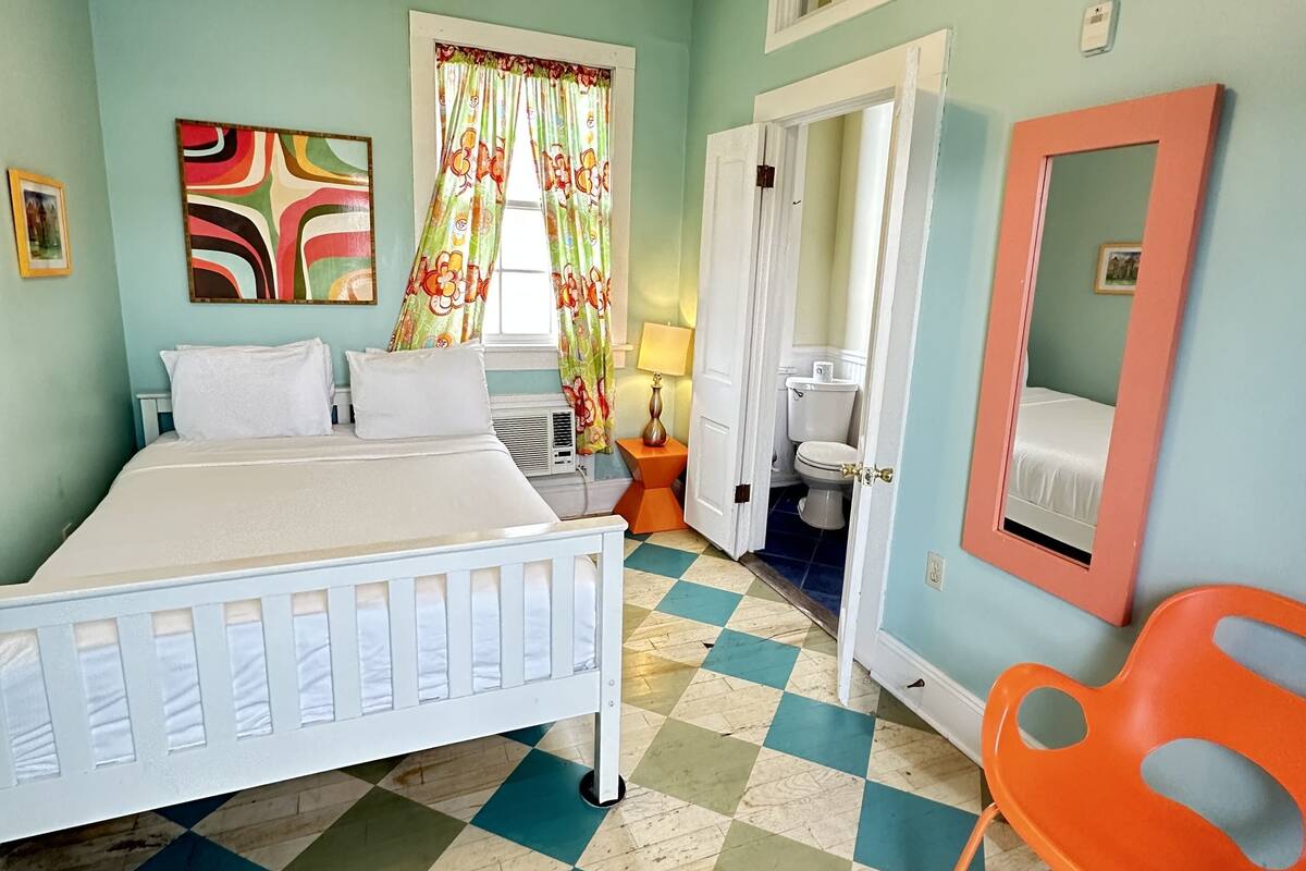 A bedroom with a bed, a ceiling fan, a window with curtains, a bathroom with a toilet, and a mirror.