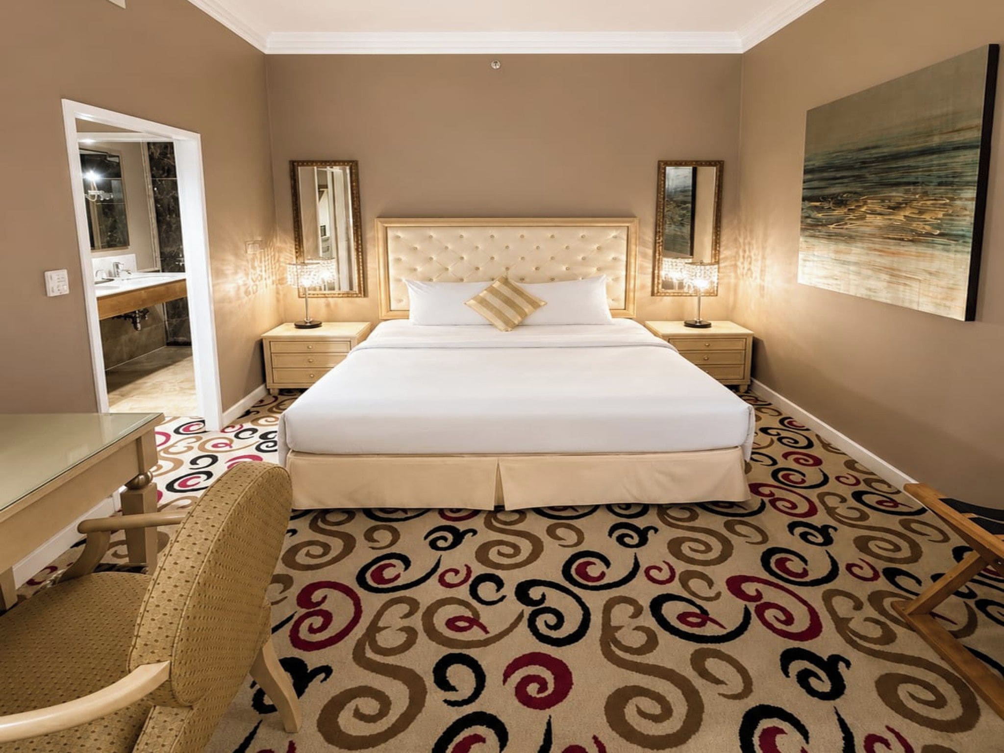 Habitación de hotel con una cama grande, dos mesitas de noche con lámparas, un escritorio, una silla y una alfombra estampada.