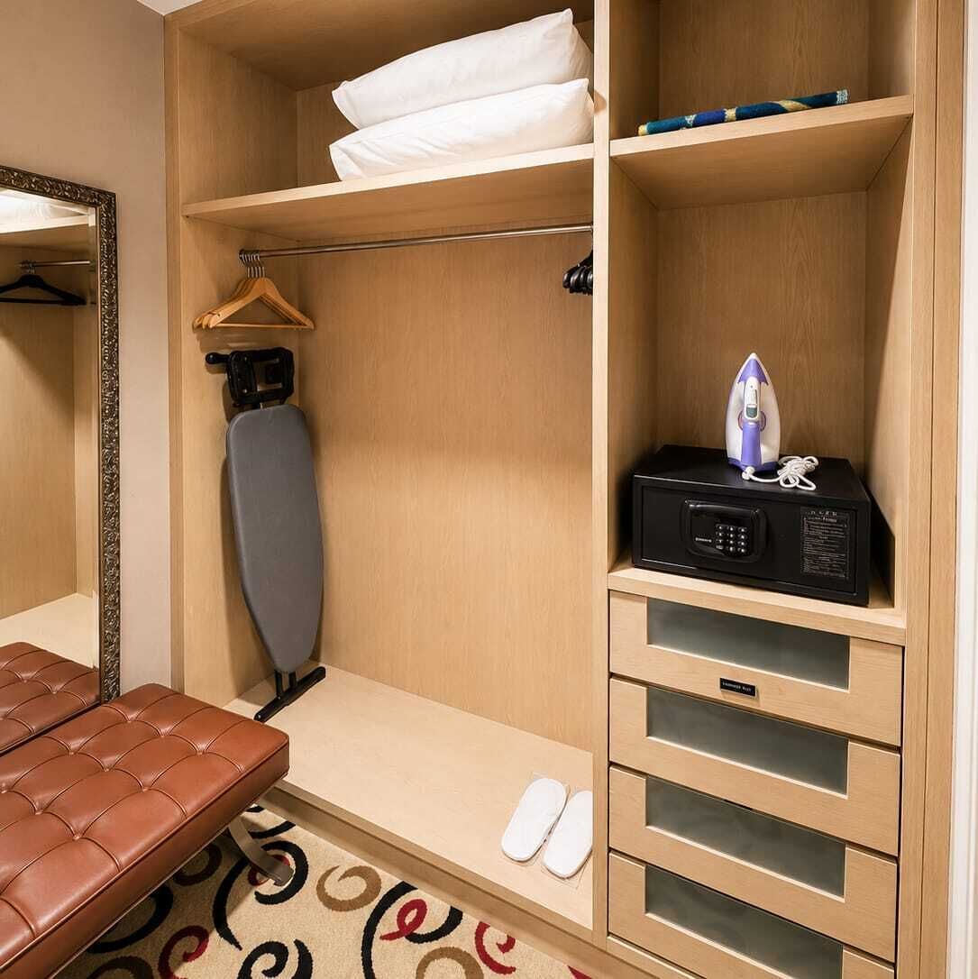 A hotel room with a bed, an ironing board, a microwave, a hanging rack, a bench, and a pair of slippers.