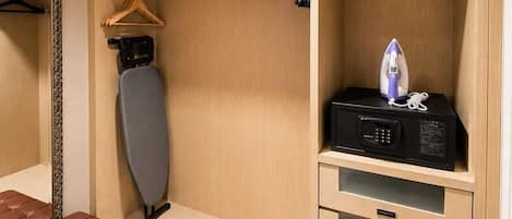 A hotel room with a bed, an ironing board, a microwave, a hanging rack, a bench, and a pair of slippers.