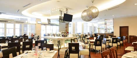 A spacious restaurant with multiple dining tables and chairs, a central bar area, and large windows allowing natural light.