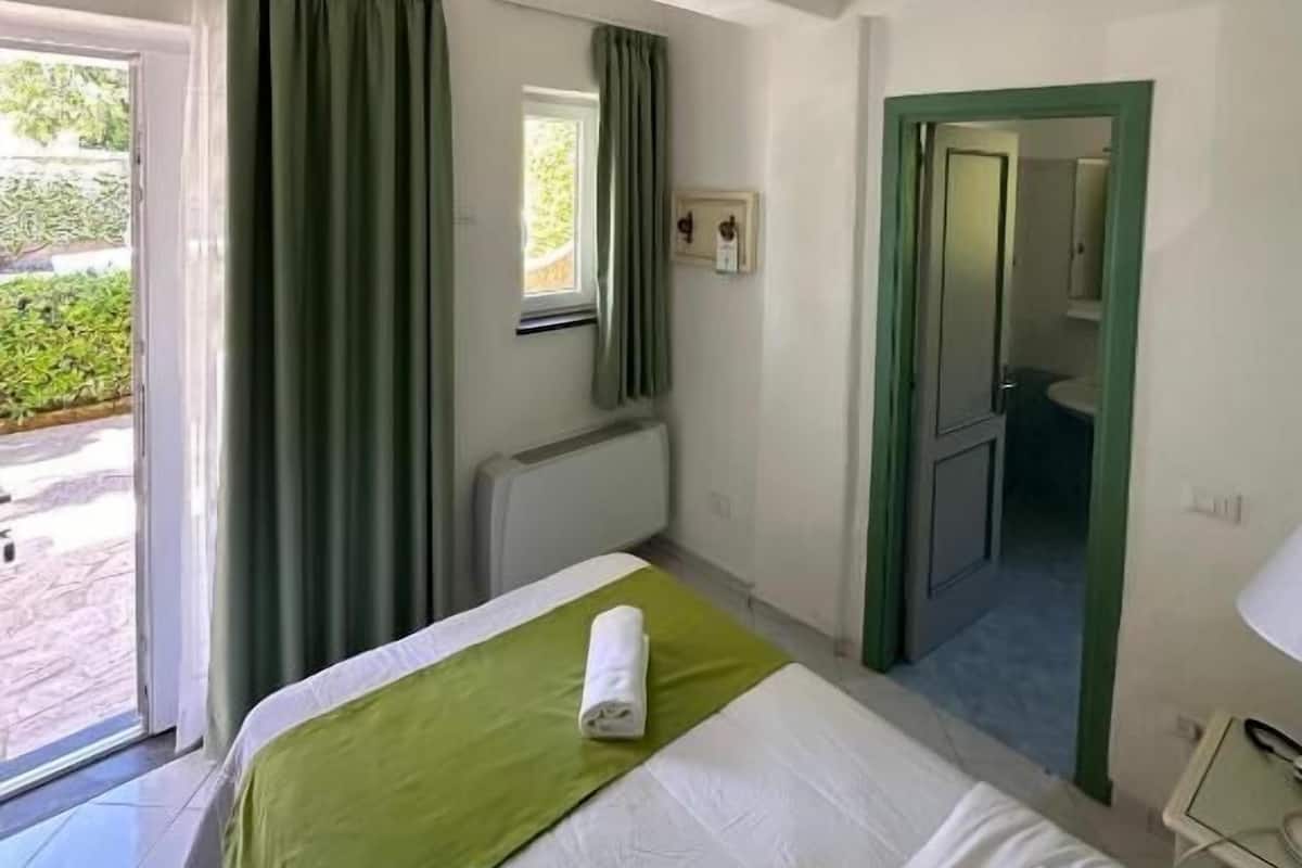A hotel room with a bed, a window with curtains, a radiator, a door, and a small bedside table.