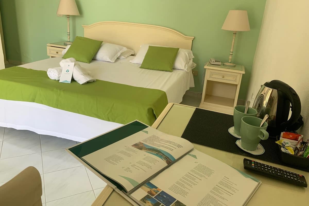 A hotel room with a neatly made bed, a desk with a remote control, a coffee cup, and a menu.