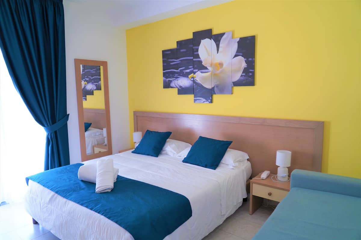 A hotel room with a bed, a nightstand, a bedside table, a mirror, and a wall with a floral painting.