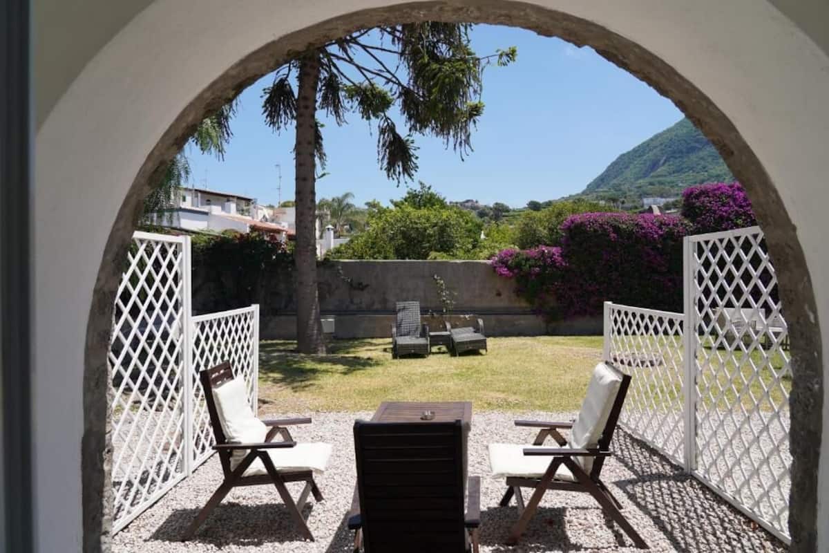 An outdoor seating area with two chairs and a table, enclosed by a white lattice fence, with a view of a mountain and greenery.