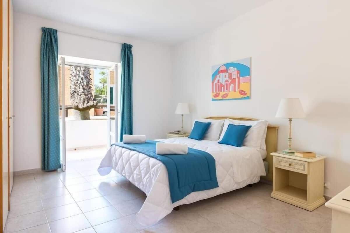 A bedroom with a bed, two bedside tables, a window with curtains, a wall painting, and a view of the outside.