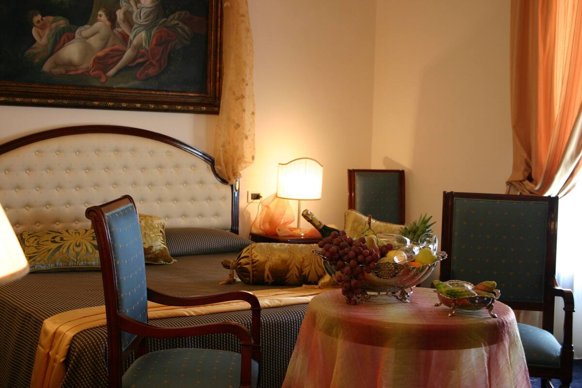 A hotel room with a bed, a table with fruit, a lamp, and a painting on the wall.