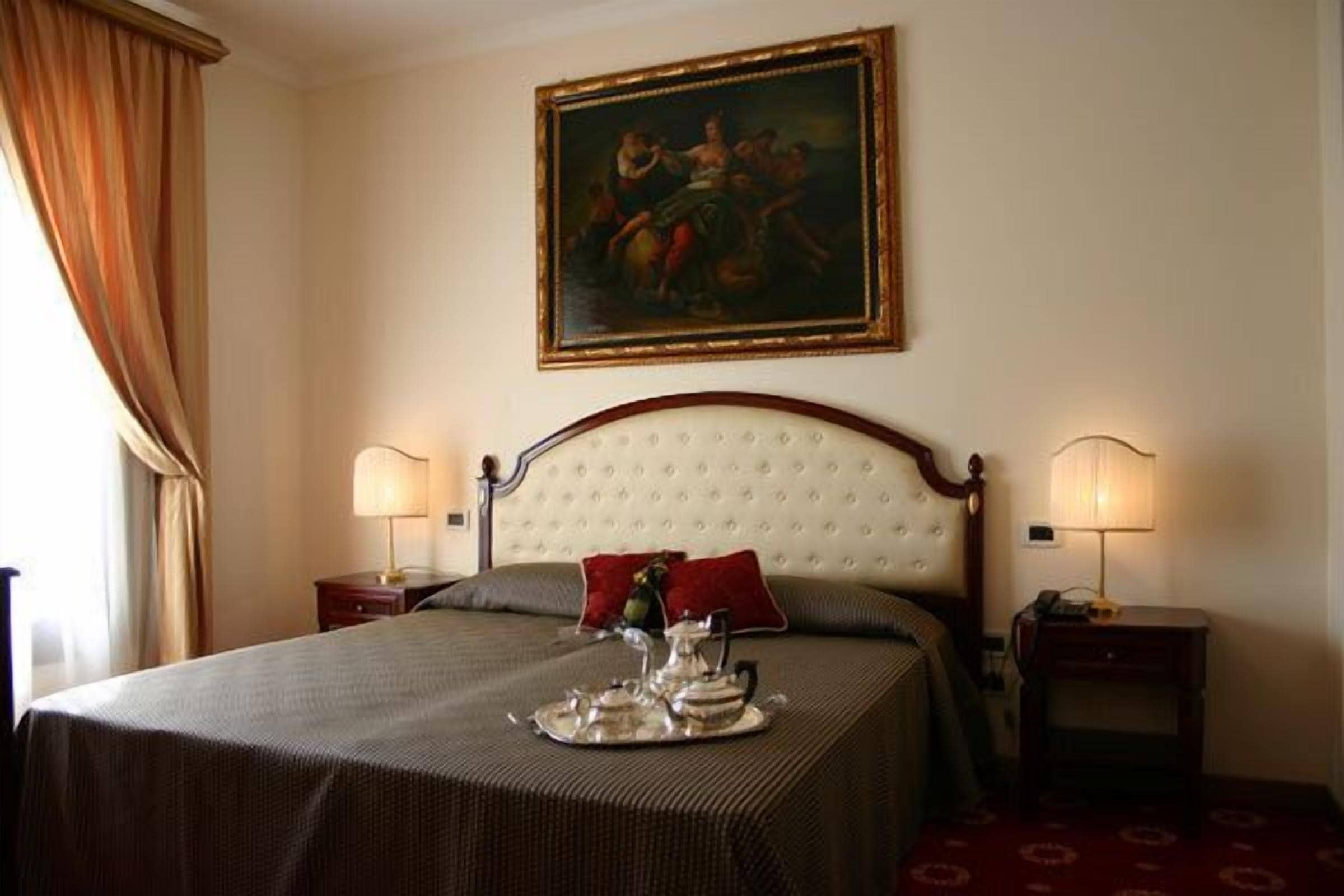A hotel room with a large bed, two bedside lamps, a painting on the wall, and a tray with a teapot and cups on the bed.