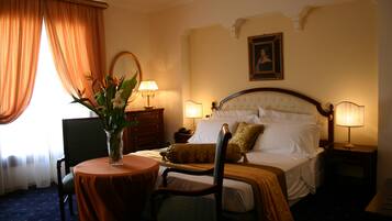 Habitación de hotel con cama, dos mesitas de noche con lámparas, una mesa redonda con una silla, cómoda y un ventanal con cortinas.