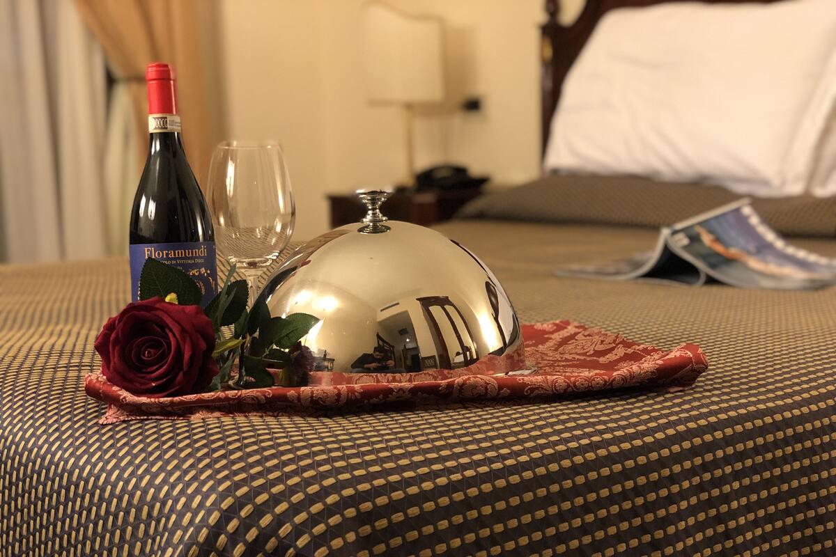 A bed with a patterned cover, a bottle of Floramundi wine, a glass, a silver tray, and a red rose on a table.