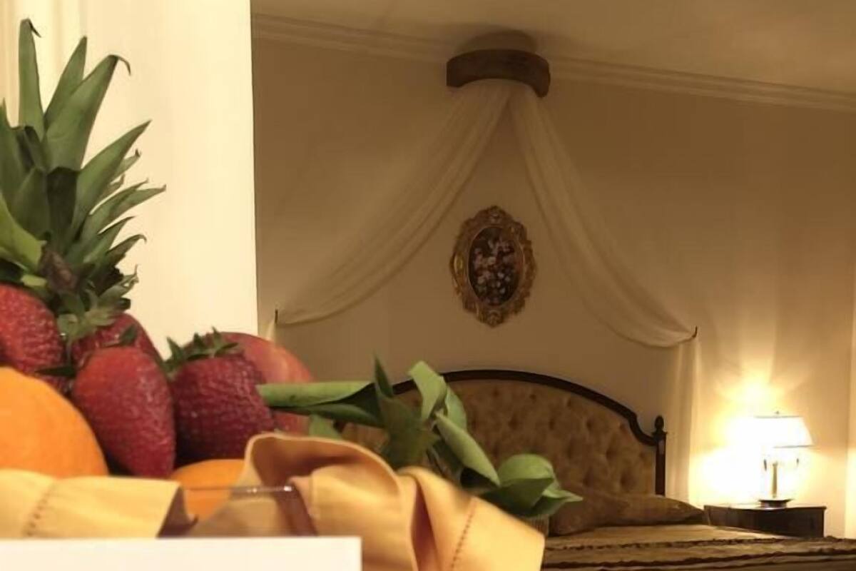 A hotel room with a bed, a bedside table with a lamp, and a table with fruit and a gift box.